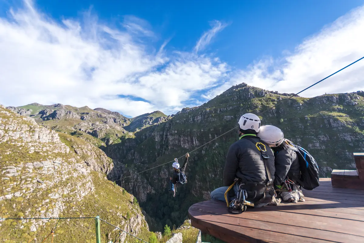 A couple sitting together on a zipline platform watching another guest fly across the Elgin Valley gorge near Cape Town